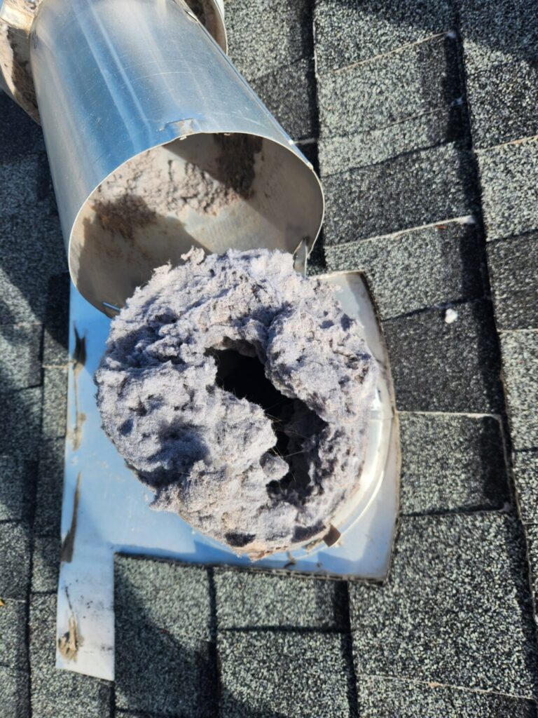 Thick gray lint buildup blocking a dryer vent opening beside the removed roof vent cap.